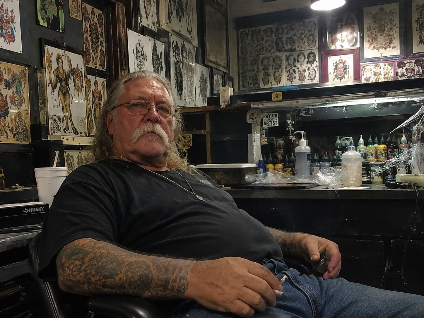 The Tattoo Shop Oracle — The Old School Artist Every Shop Still Needs Old school tattoo artist seated inside a tattoo shop surrounded by flash sheets and tattoo equipment
